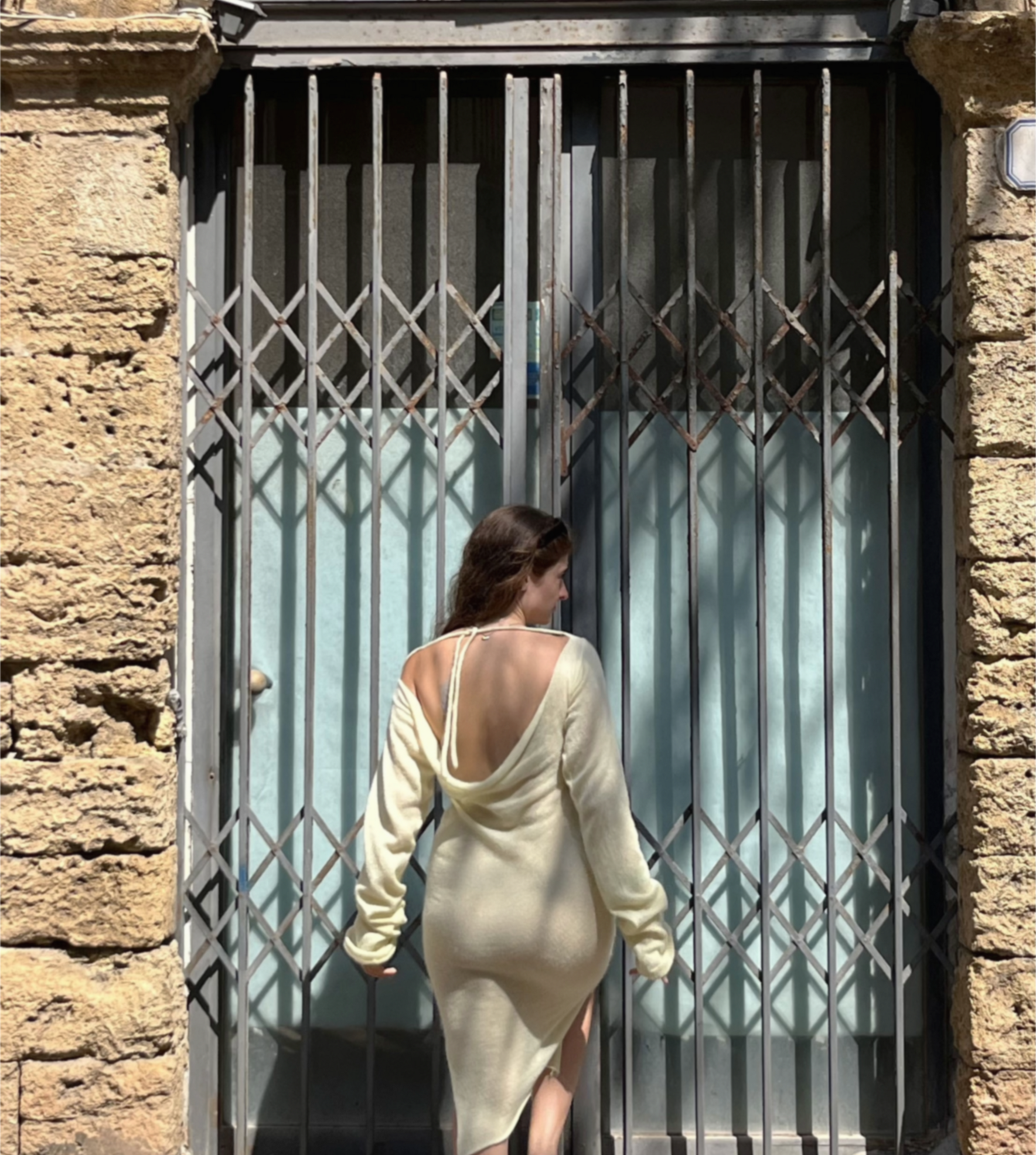 A woman stands with her back to the camera in front of a metal security gate, wearing a cream-colored, long-sleeve, open-back knit dress.